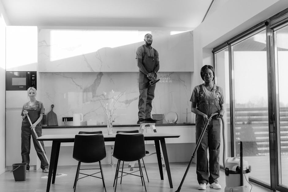 A woman with long curly hair, dressed in a beige jacket, blue jeans, and white sneakers, is engaged in domestic cleaning in a modern kitchen. She is sweeping the floor with a broom, focusing on the wo